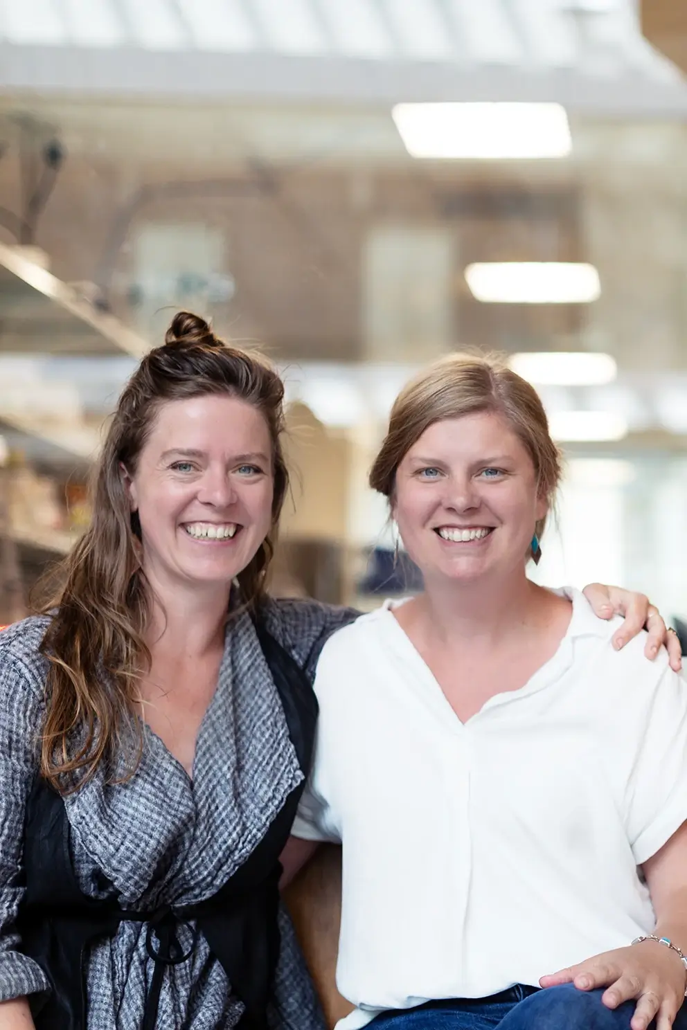 Two smiling women with arms around each other in modern office setting with bright lighting.