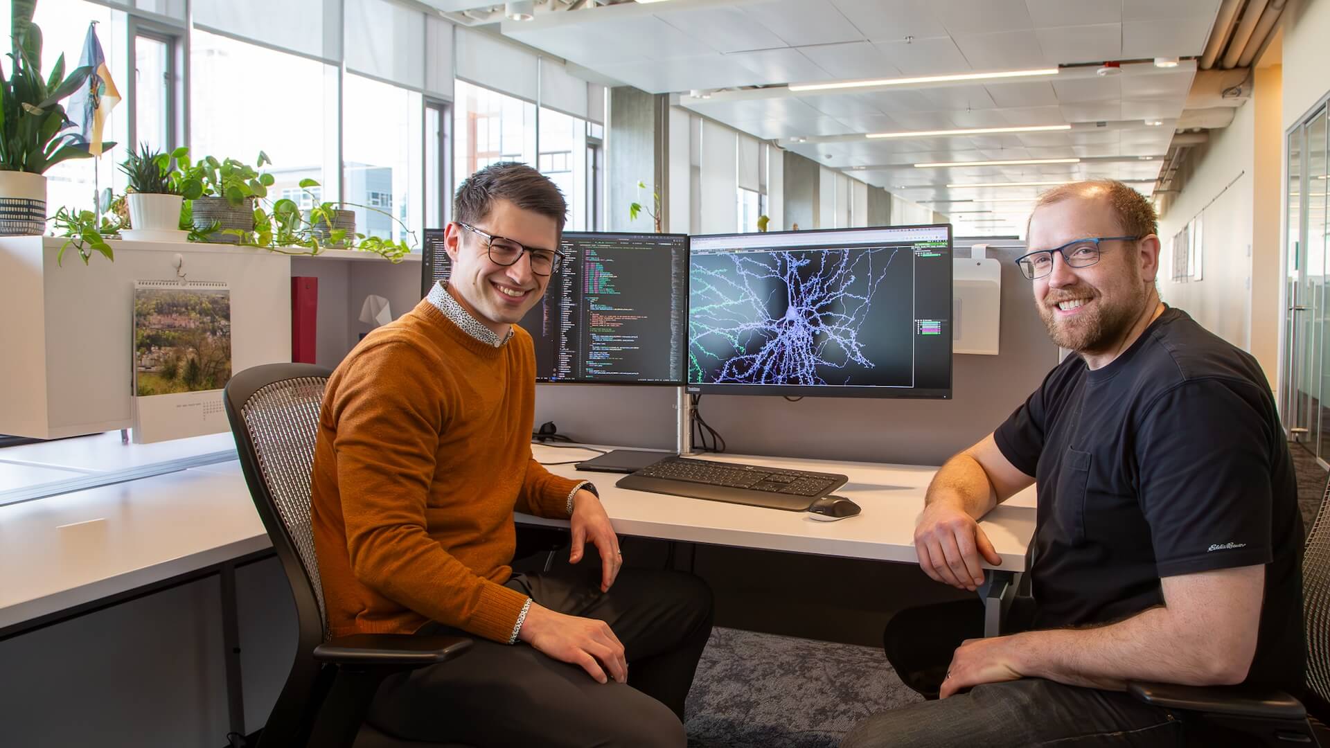 Two smiling men in modern office with code monitors and plants visible behind them.