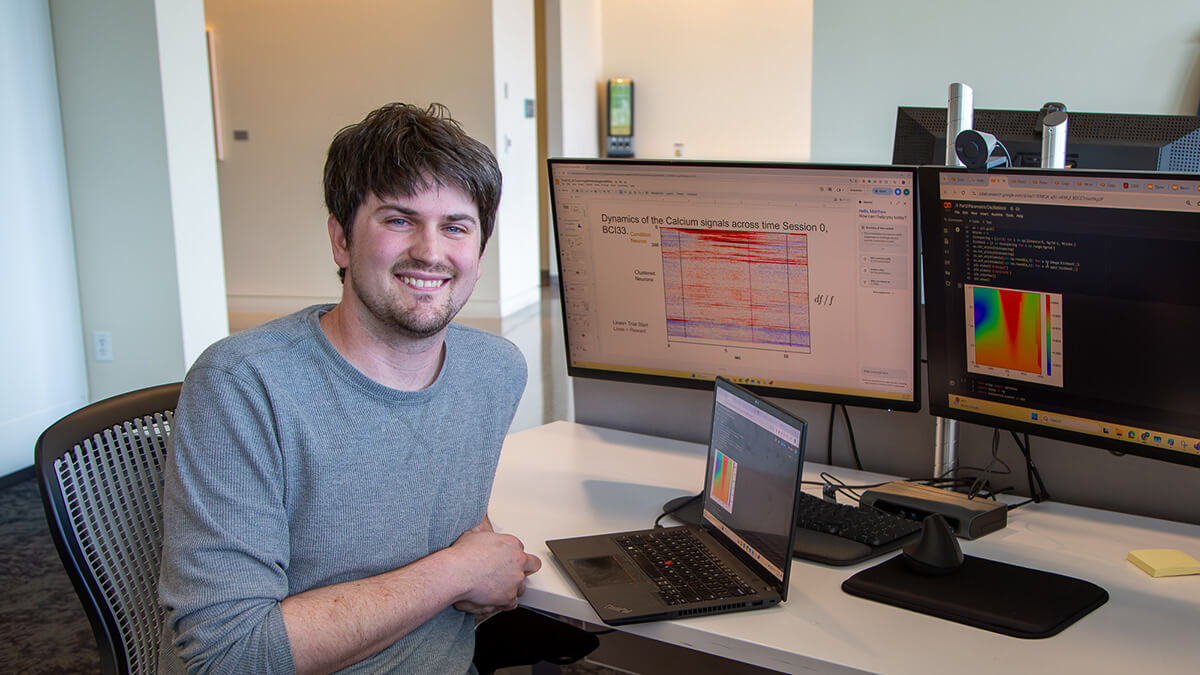 Man smiling at desk with multiple computer monitors displaying scientific data visualizations