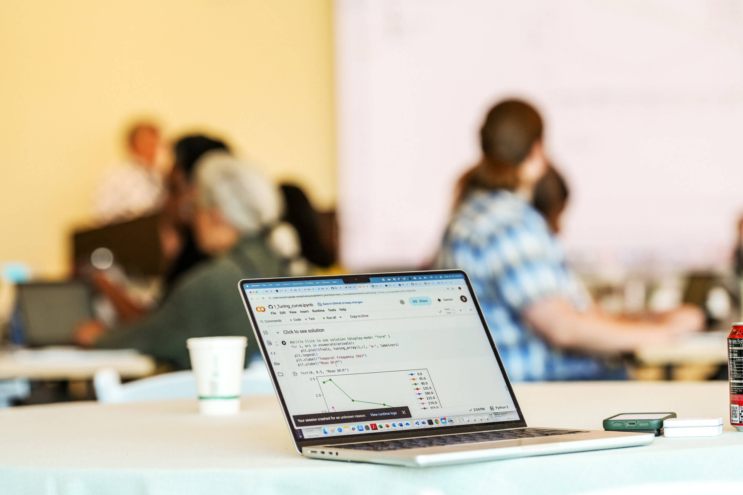 Laptop displaying code on desk with blurred people collaborating in background office setting.