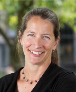Woman smiling at camera, wearing black blazer and beaded necklace outdoors