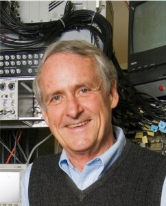 Older man smiling in front of electronic equipment and vintage computer monitors.