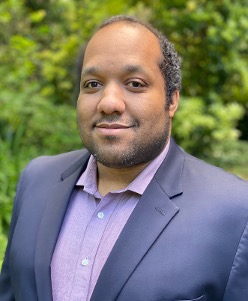 Professional portrait of a man wearing a light purple shirt and navy blazer, smiling outdoors with green foliage background