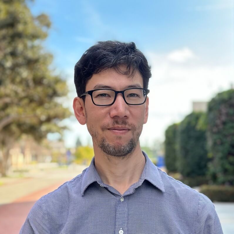 Man with glasses wearing blue shirt smiling at camera outdoors