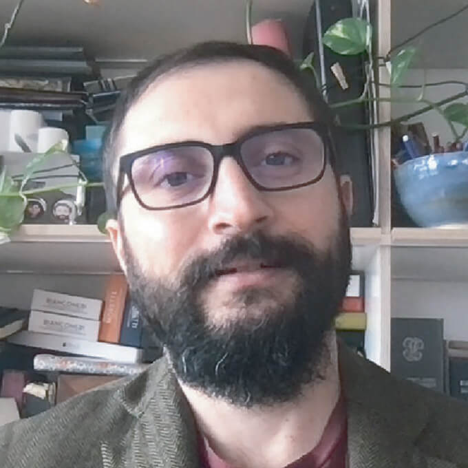 Man wearing glasses and beard posing indoors with shelves of books and plants behind him