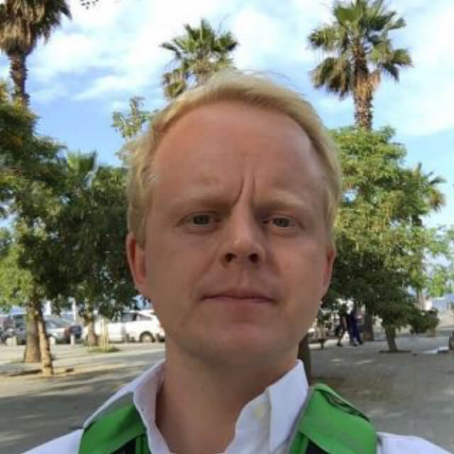 Man in green and white jacket outdoors with palm trees and buildings in background