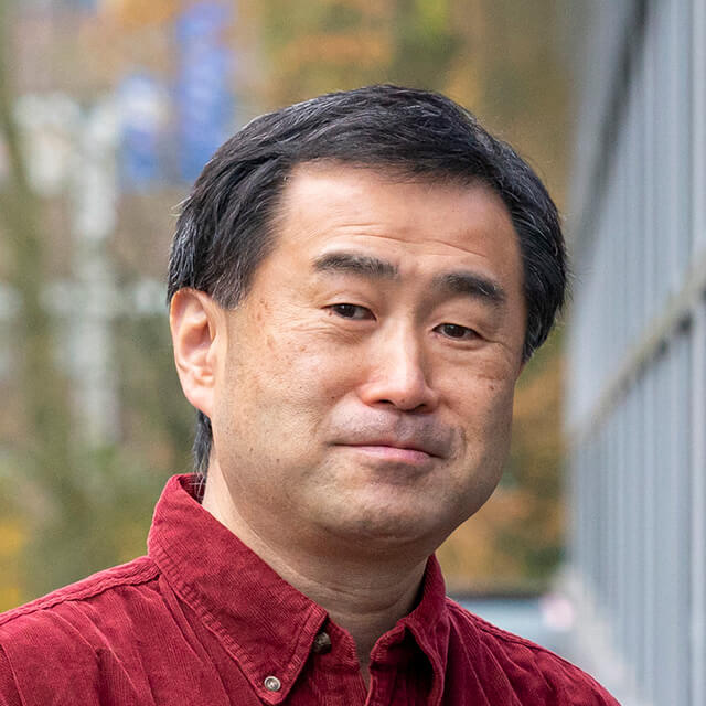 Middle-aged Asian man in red shirt, outdoor portrait with blurred garden background.