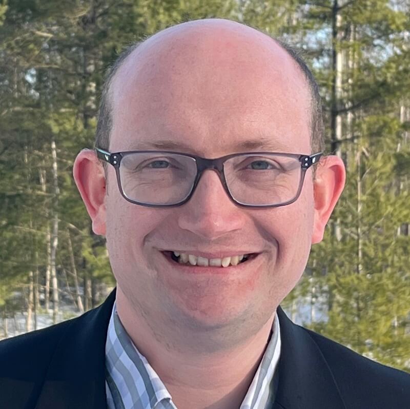 Bald man wearing glasses and dark blazer smiles outdoors with forest background