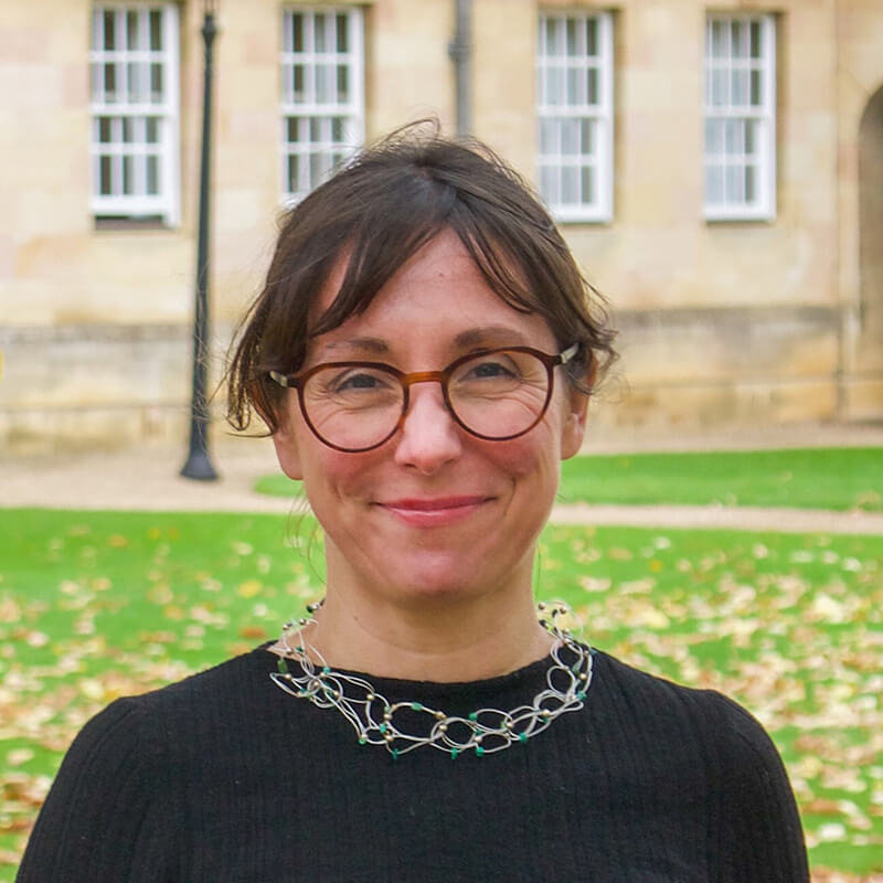 Person with glasses and chain necklace smiling outdoors near historic building