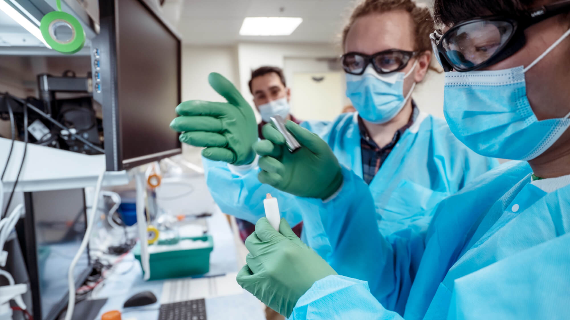 Scientists in lab coats and masks examining samples with green gloves in laboratory setting.