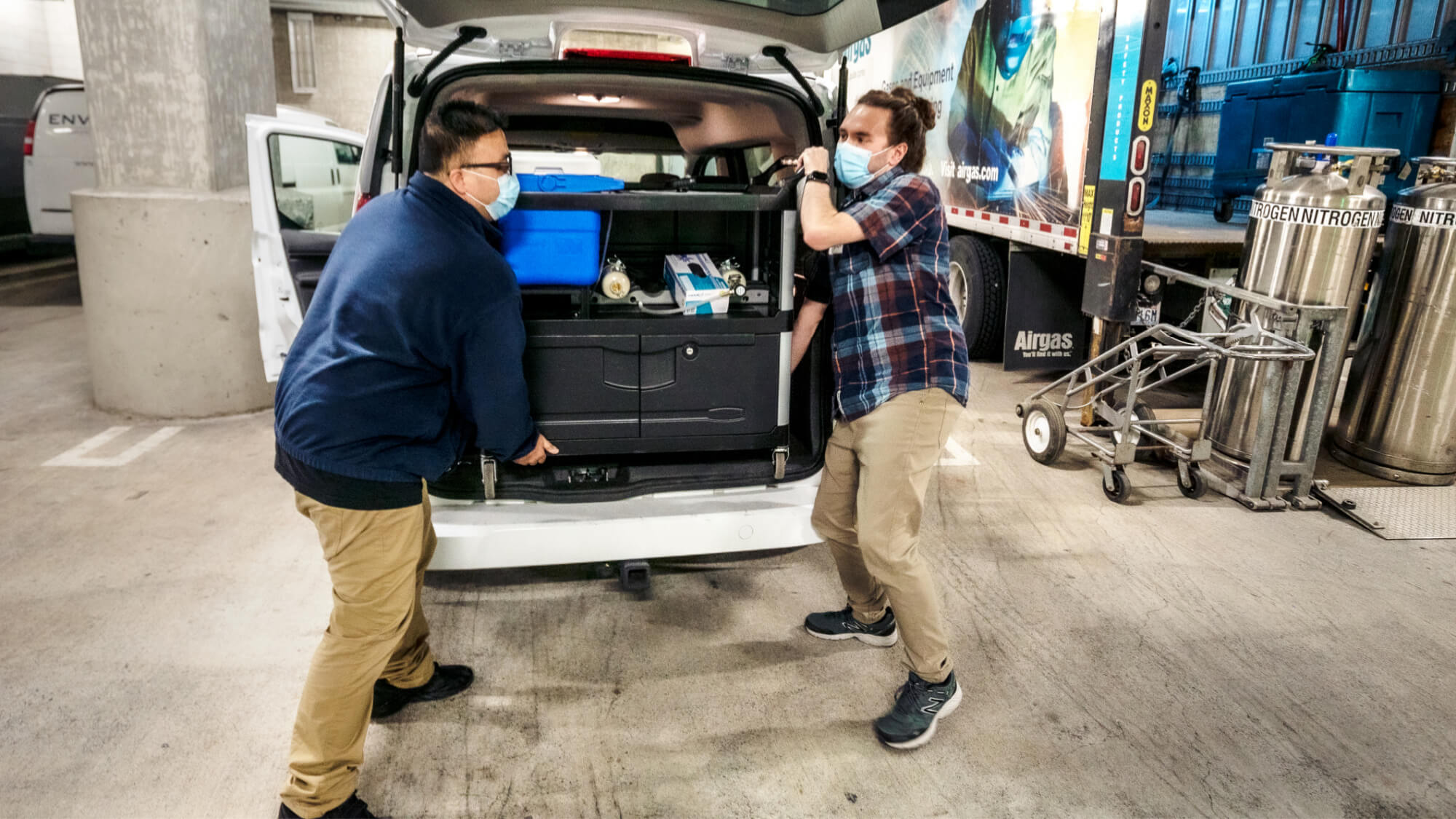 Two masked workers unloading equipment from open van cargo area in industrial warehouse facility.