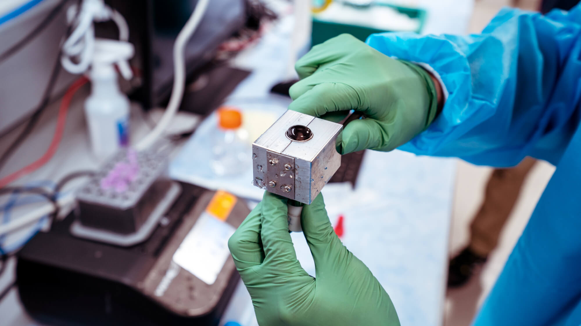 Scientist in green gloves holds metal component in laboratory setting with electronics.