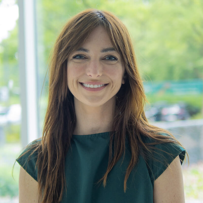 Woman with long brown hair smiling at camera near bright window