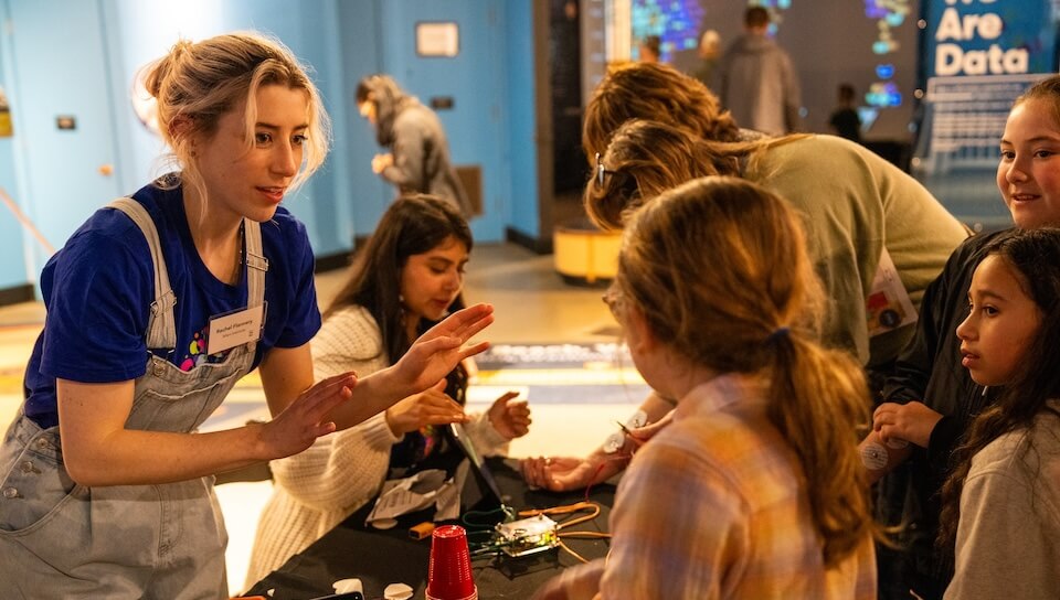 Museum educator in overalls demonstrates interactive science exhibit to engaged children indoors.