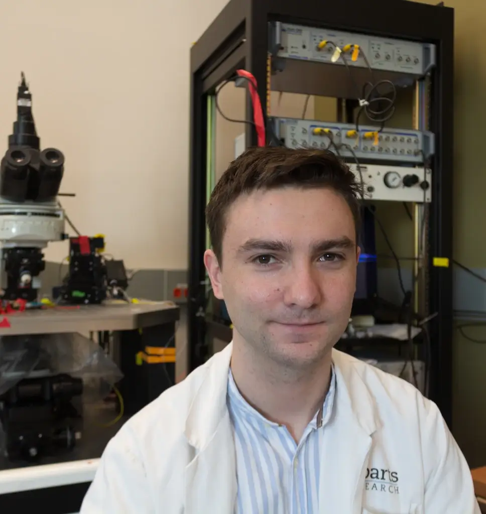Man in lab coat smiles in laboratory with scientific equipment and 3D printer visible.
