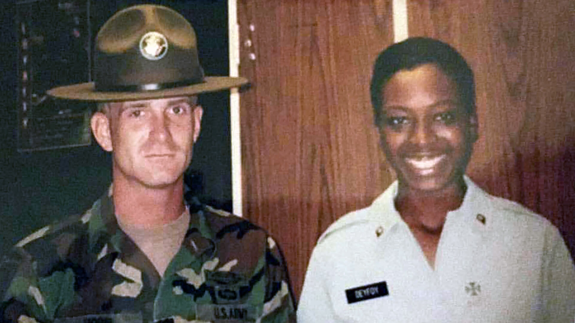 Military drill sergeant in campaign hat standing next to smiling service member indoors