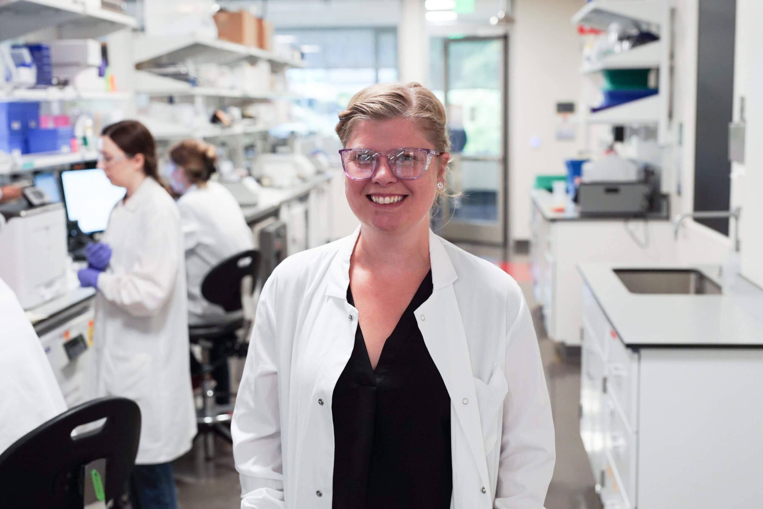 Smiling scientist in white lab coat and glasses standing in modern laboratory with colleagues working in background