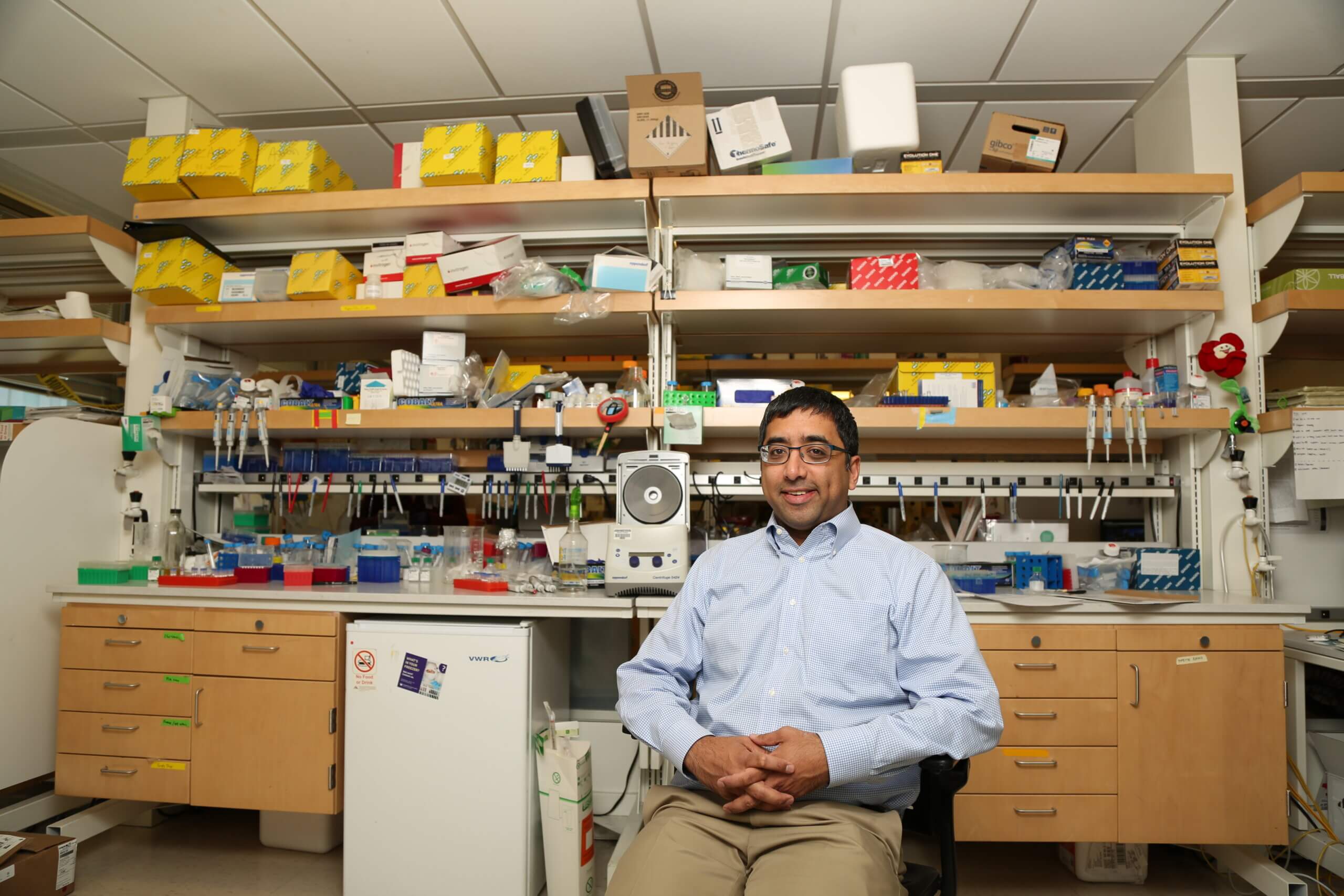 Man in glasses sitting in electronics lab with shelves of components and equipment.