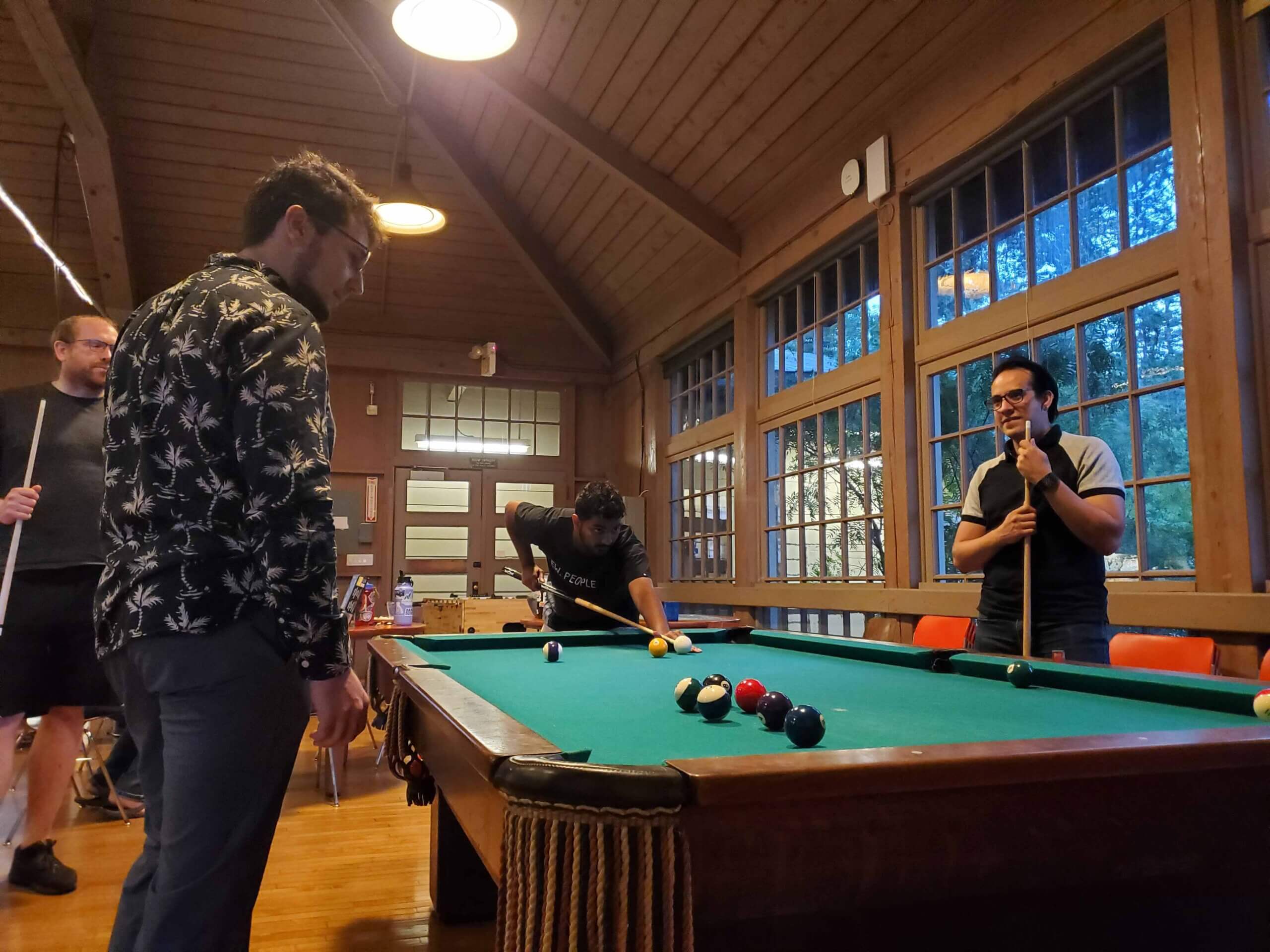 Four men playing pool billiards in wooden cabin with large windows at night.