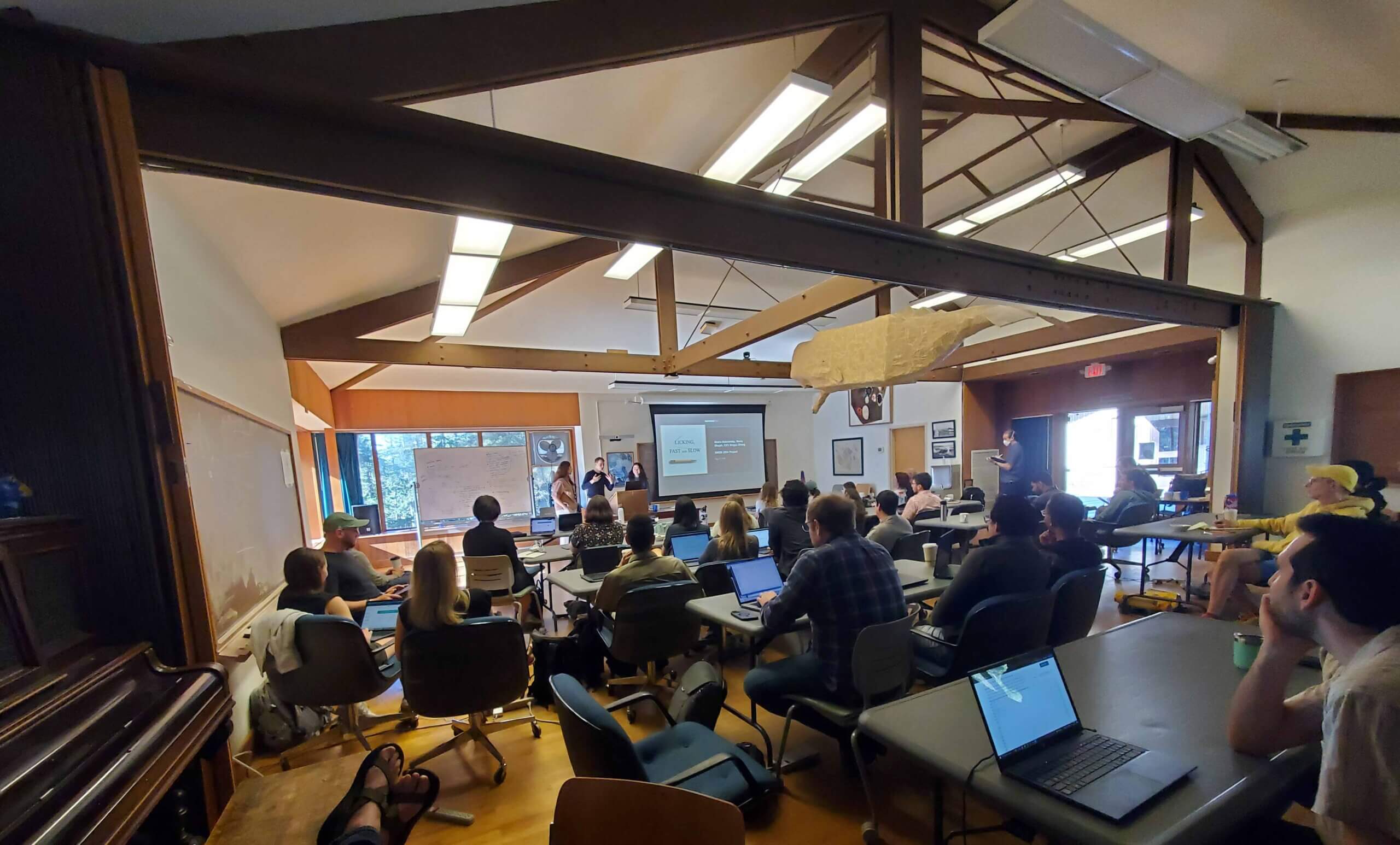 Large classroom with students seated at tables with laptops, listening to presenter near projection screen