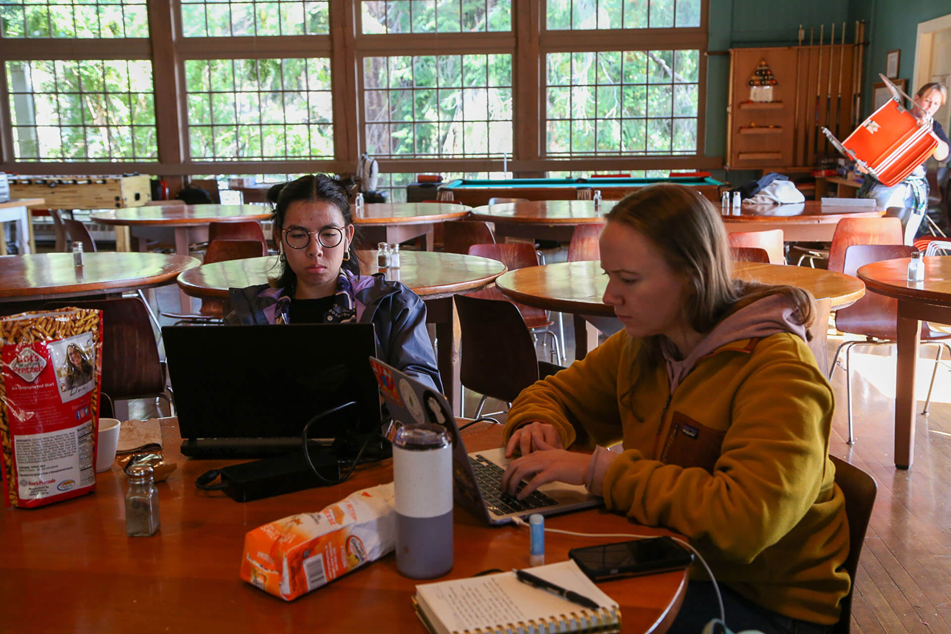 Two students studying at cafeteria table with laptops in bright classroom with large windows