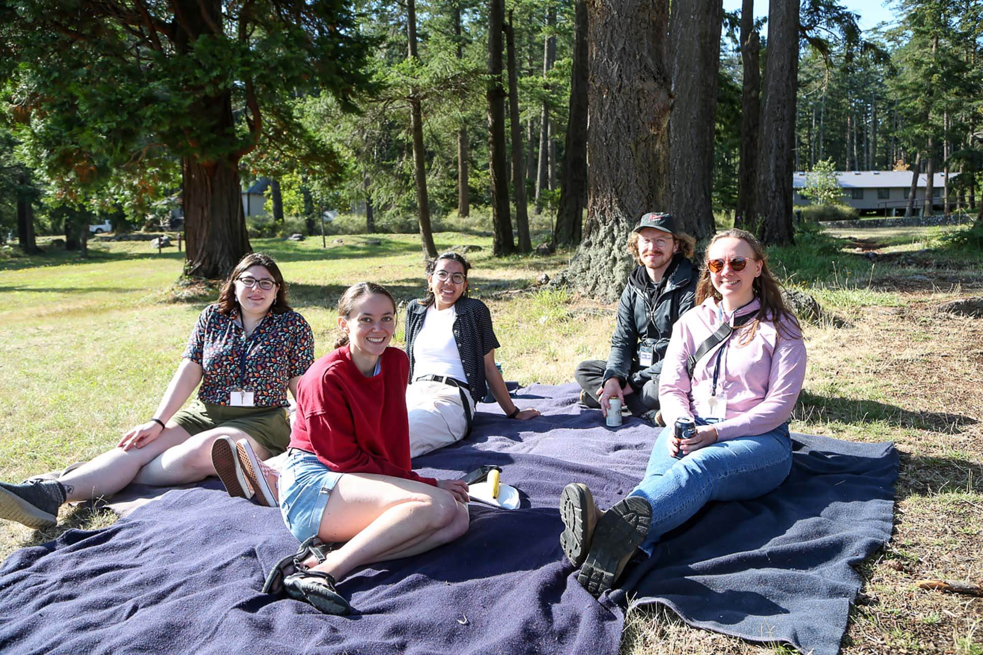 Five smiling people sitting on a blanket in a forest picnic area with tall trees