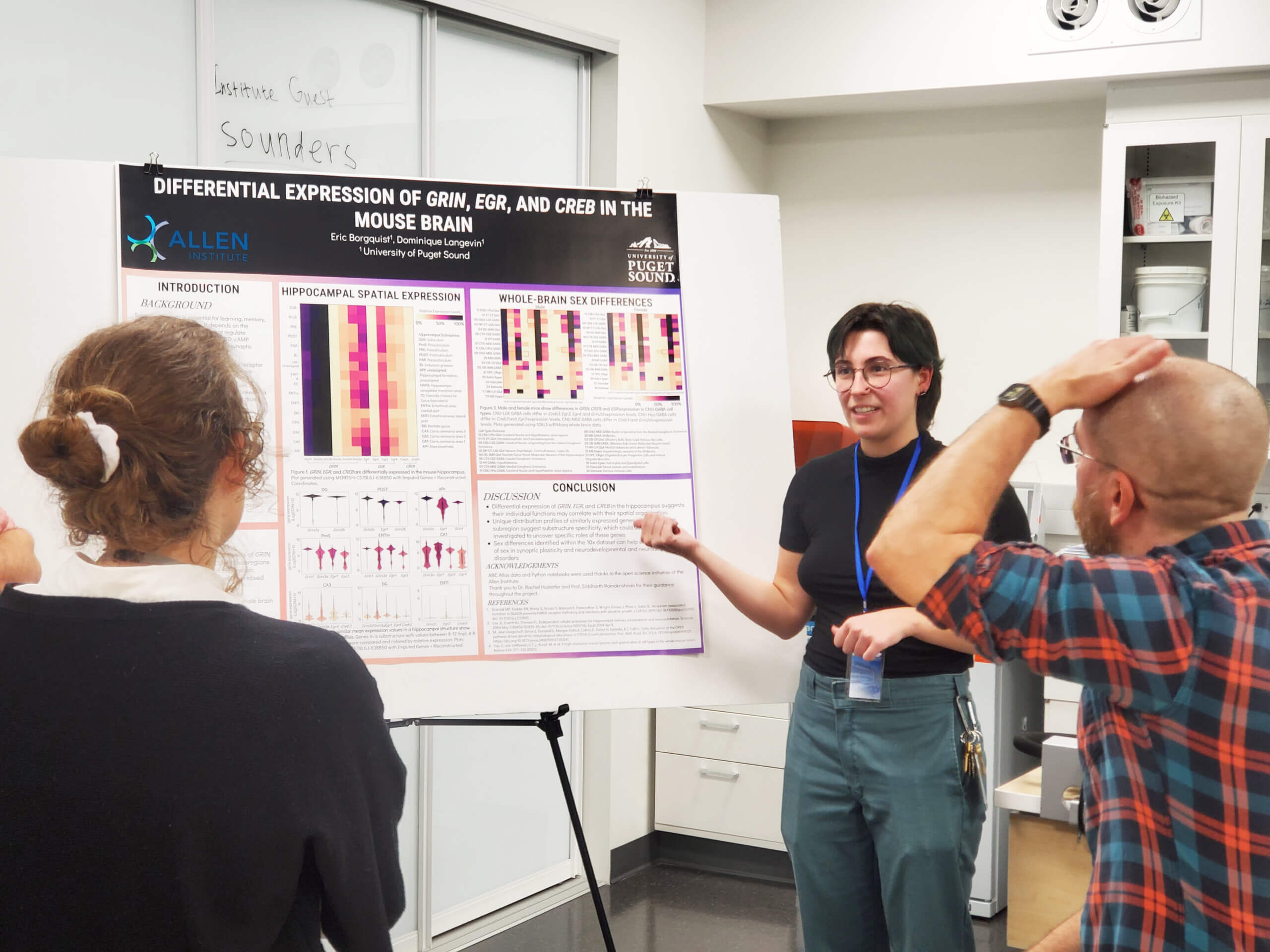 Researcher presenting neuroscience poster on brain gene expression to two attendees at academic conference.