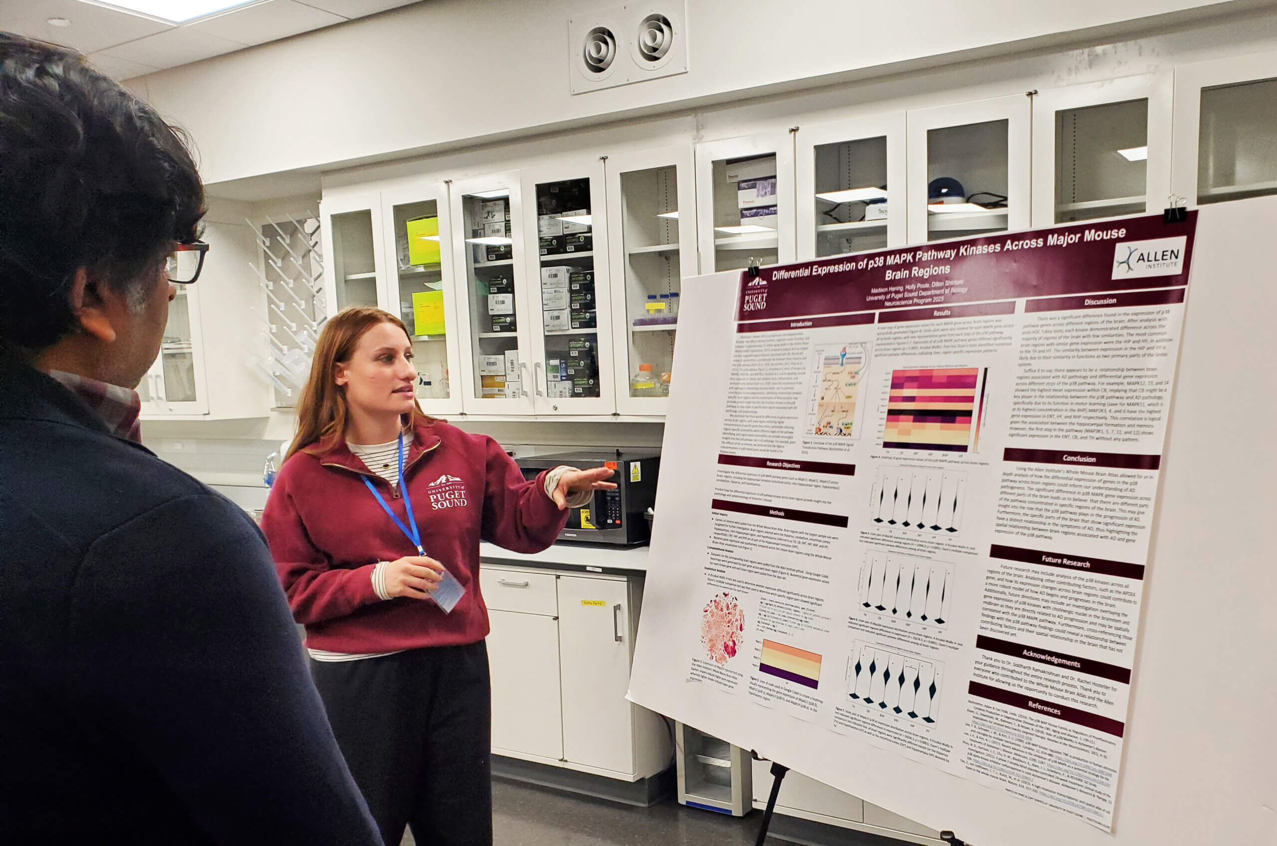 Researcher presents scientific poster on MAPK pathway kinases in mouse brain to visiting attendee in lab.