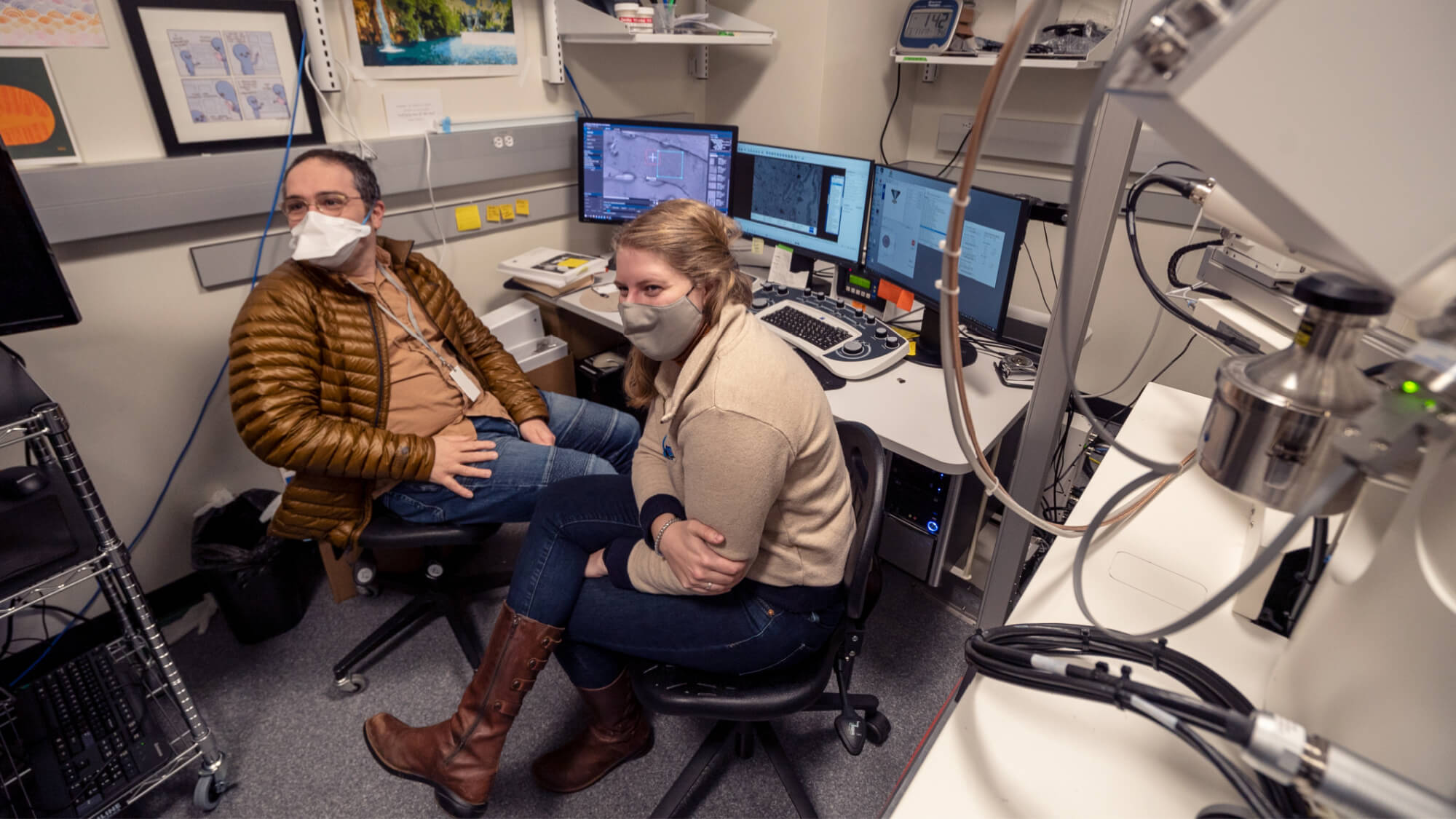 Two masked people in a high-tech laboratory with multiple computer monitors and scientific equipment