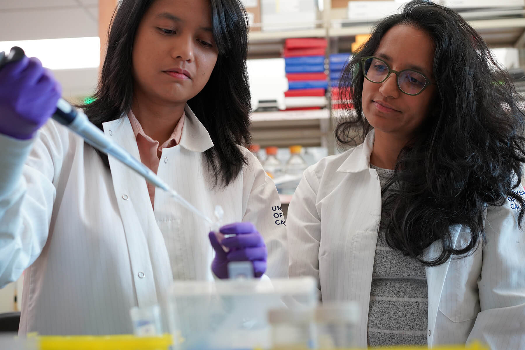 Two female scientists in lab coats conducting experiment with purple gloves and pipette.