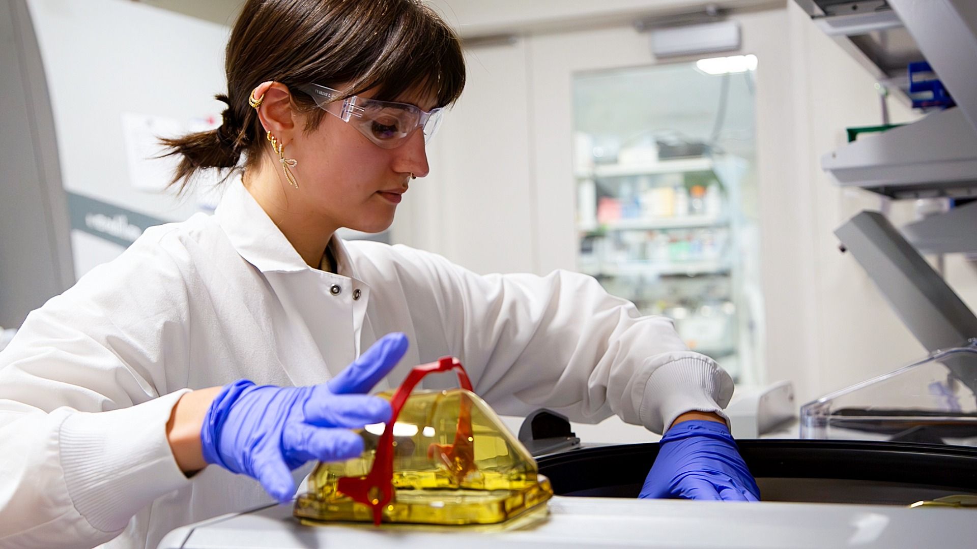 Female scientist in lab coat and safety glasses working with yellow chemical container