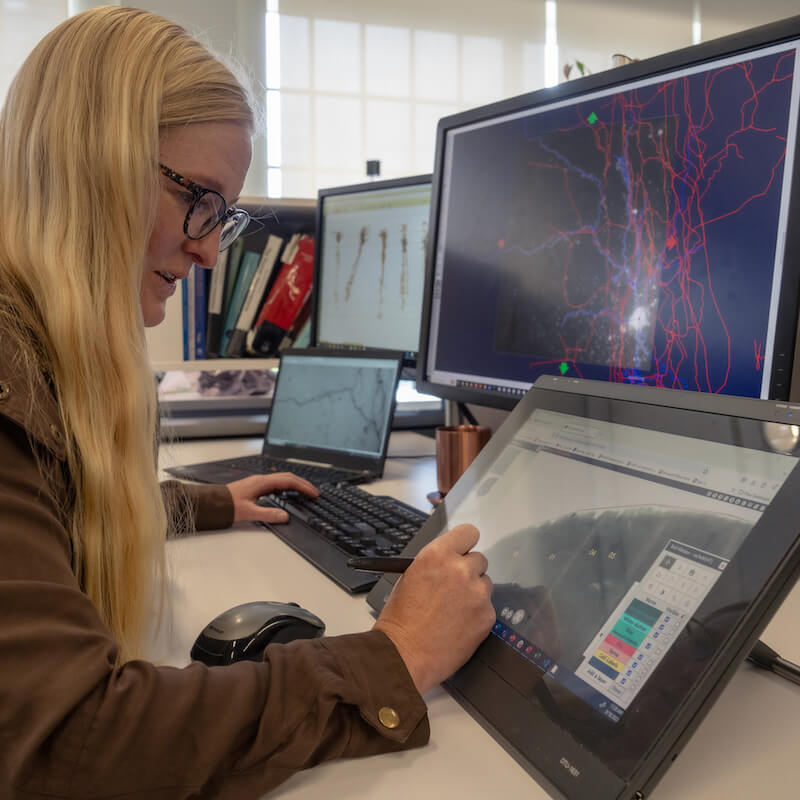 Woman with blonde hair and glasses using touchscreen tablet at desk with multiple computer monitors displaying data visualizations.