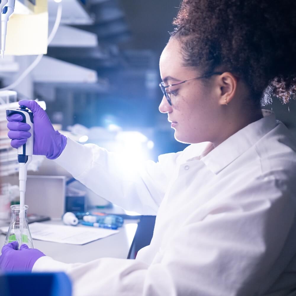 Scientist in white lab coat and purple gloves conducting laboratory experiment with pipette.