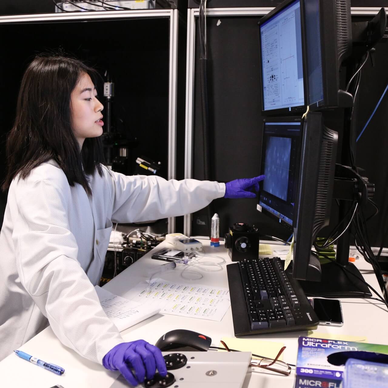 Lab technician wearing purple gloves working at computer station with multiple monitors in laboratory.