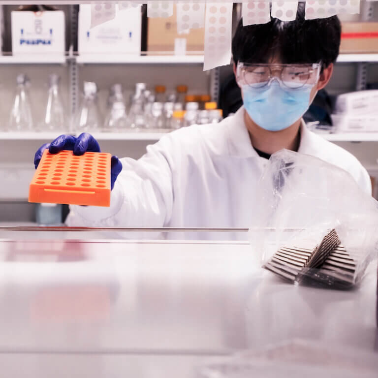 Laboratory scientist in white coat and mask holds orange sample tray in sterile lab environment