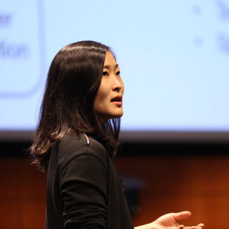 Woman with long dark hair speaking on stage with blue projection screen behind