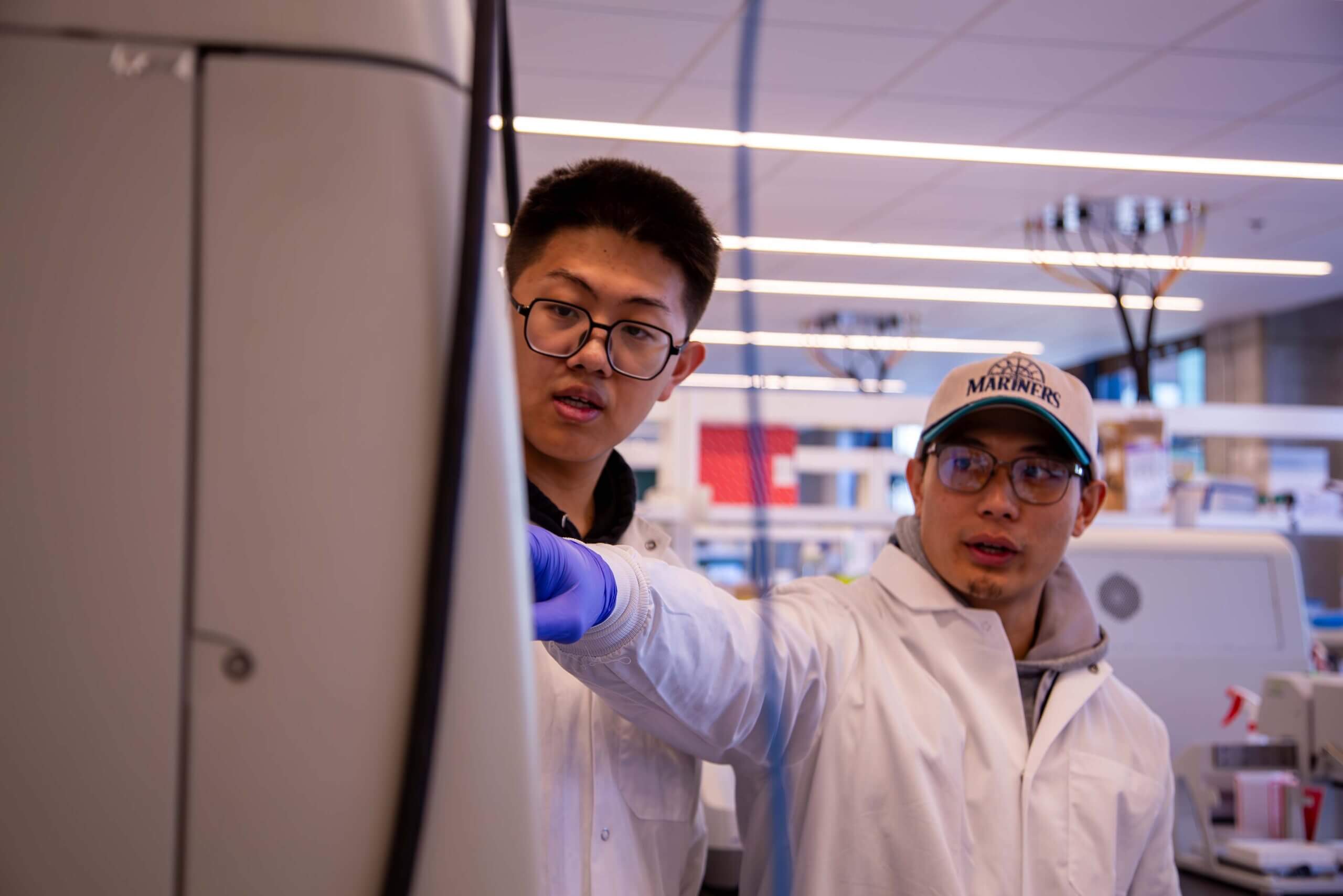 Two young scientists in white lab coats working in a modern laboratory with glowing ceiling lights.