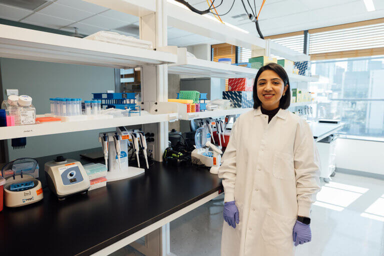 Female scientist in white lab coat smiling in modern laboratory with equipment and storage shelves.