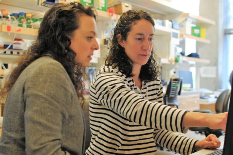 Two women examining scientific equipment in a laboratory with shelves of supplies.