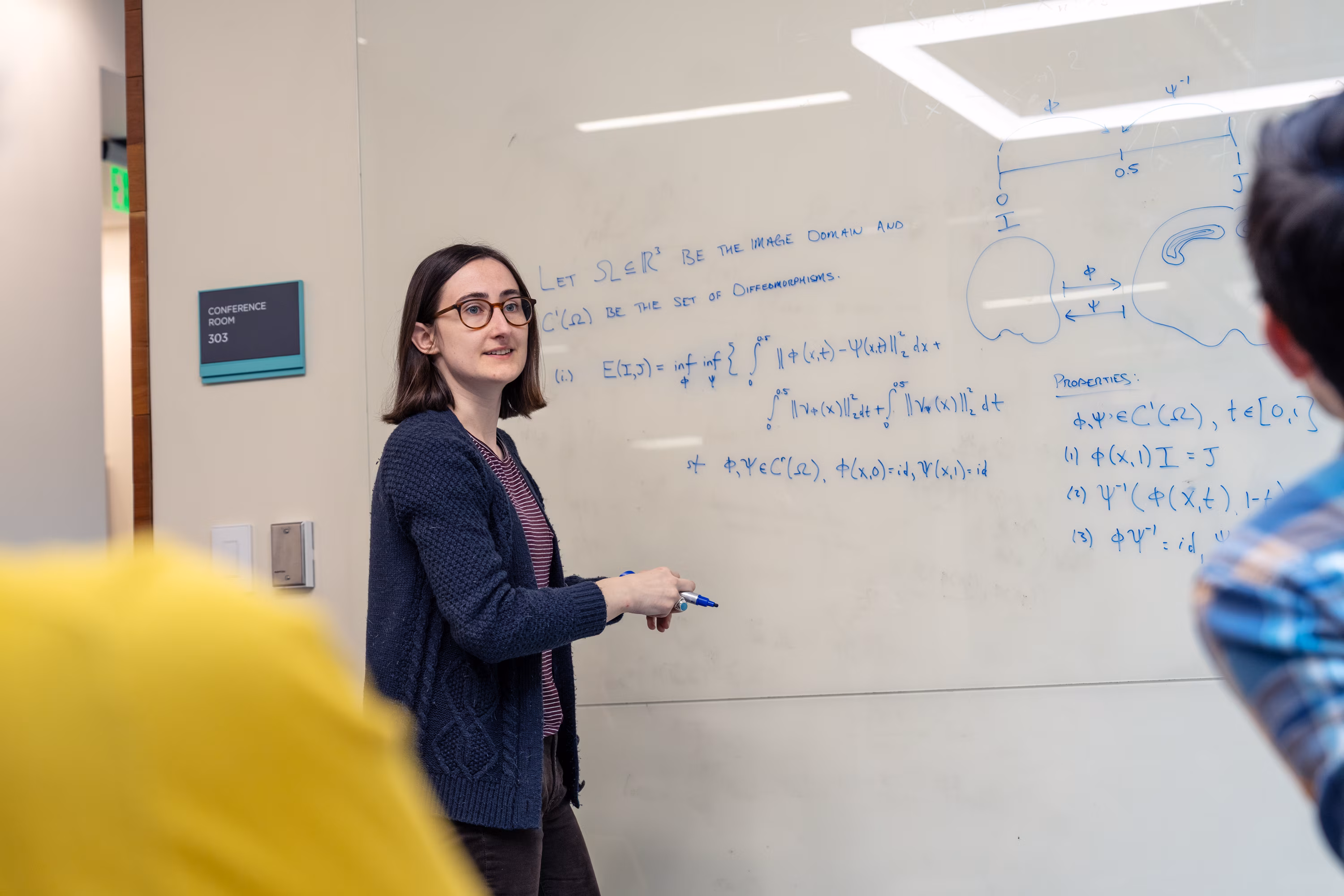 Woman wearing glasses presenting mathematical equations on whiteboard in conference room.