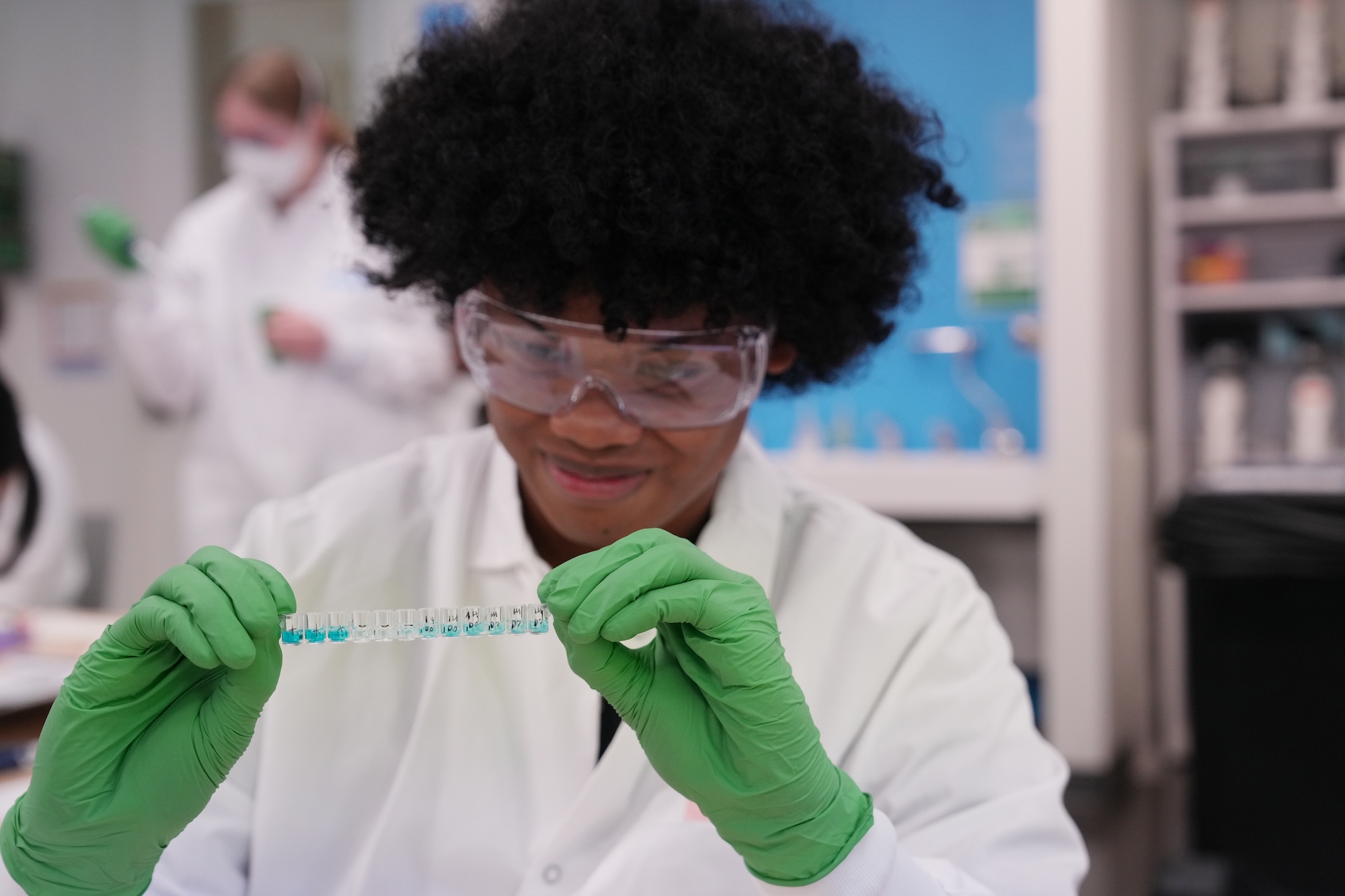 Student in white lab coat and green gloves examines DNA test samples in laboratory.
