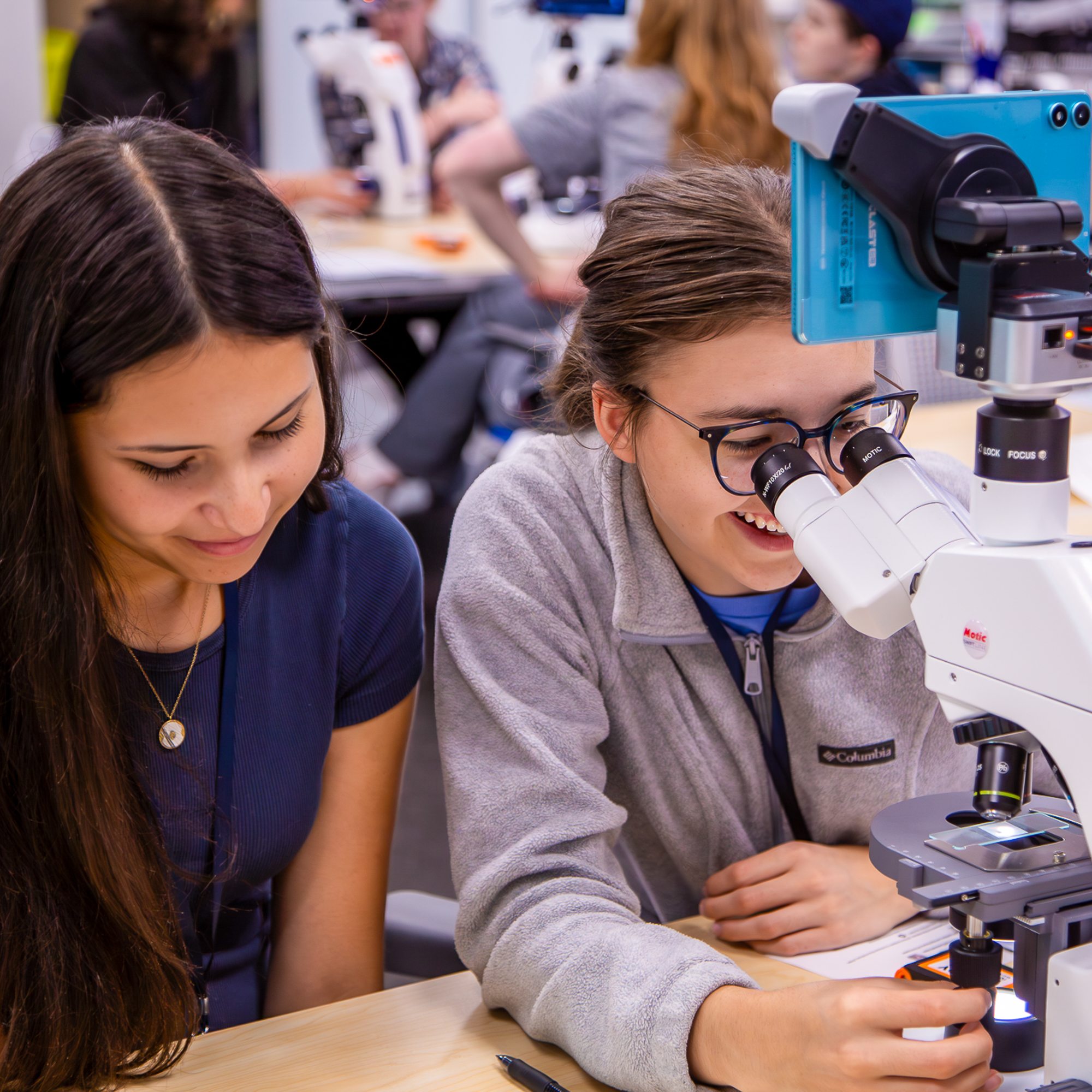 Young student examines specimen under microscope while instructor observes in science classroom laboratory.