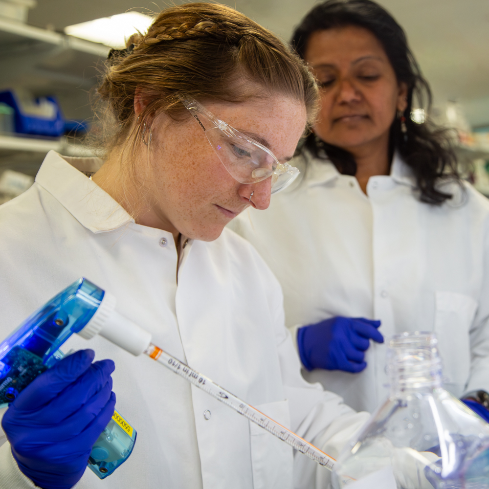 Female scientist in lab coat and safety goggles conducts research with blue gloves in laboratory.