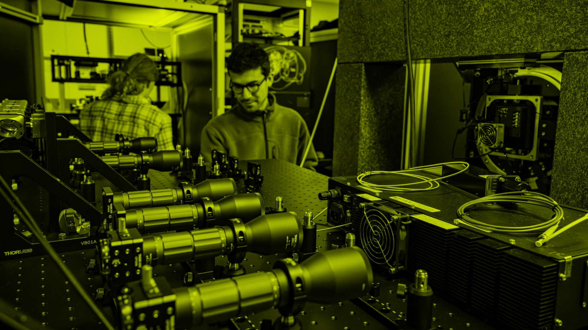 Scientists work with laser and optical equipment in a yellow-lit laboratory setting.