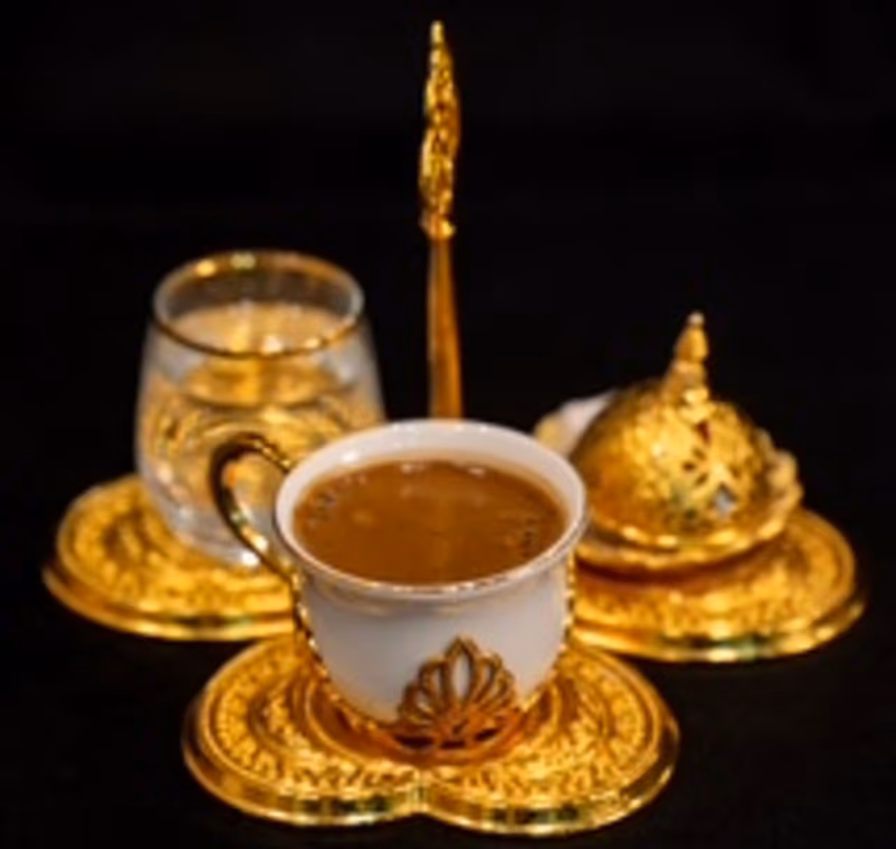 White porcelain coffee cup with gold detailing filled with Turkish coffee, accompanied by a glass of water and a covered golden sugar bowl on ornate golden trays.