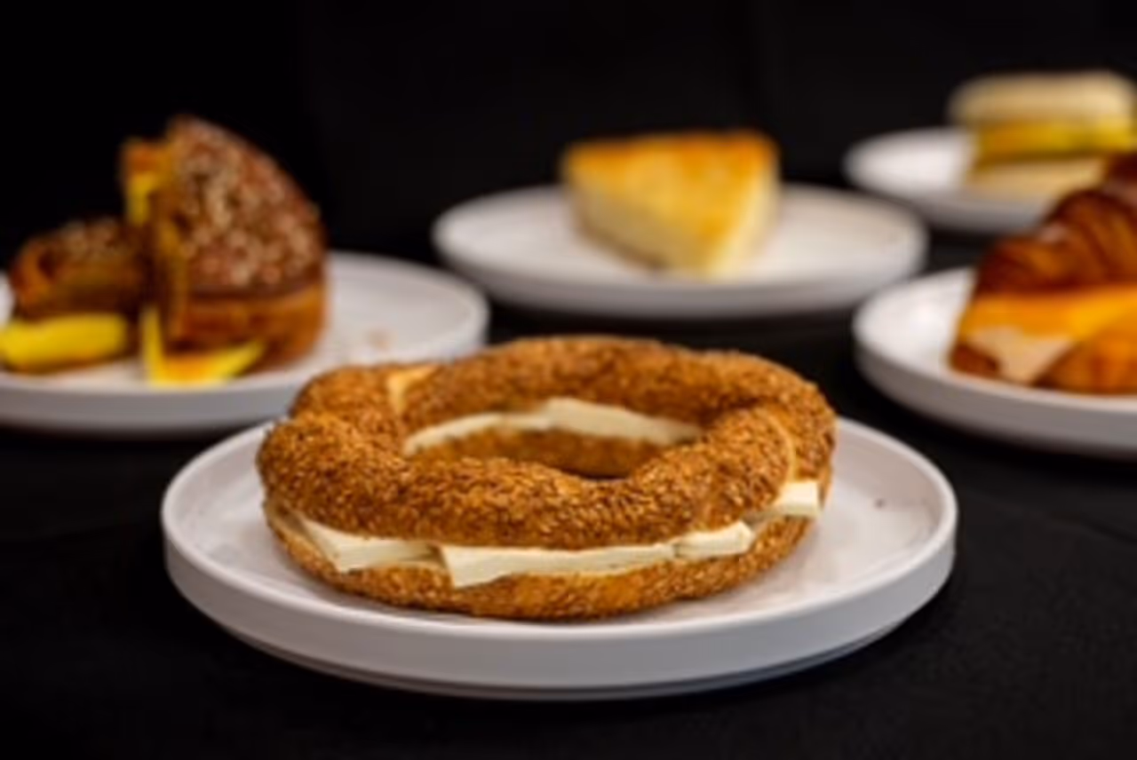 Sesame-covered bagel sandwich with cream cheese on a white plate, with other assorted sandwiches blurred in the background.