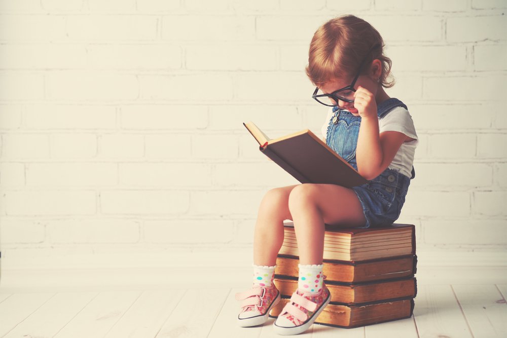 A child sits on a large stack of books reading while wearing large glasses