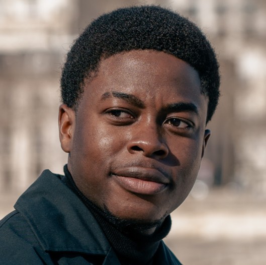 Portrait of a young Black man wearing a white button-up shirt against a gray background.
