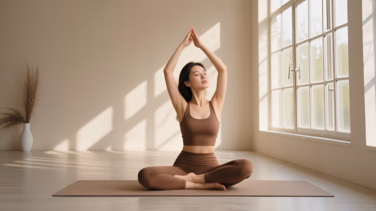 Woman in brown activewear seated cross-legged on a yoga mat with arms raised above her head in a bright room near a large window.