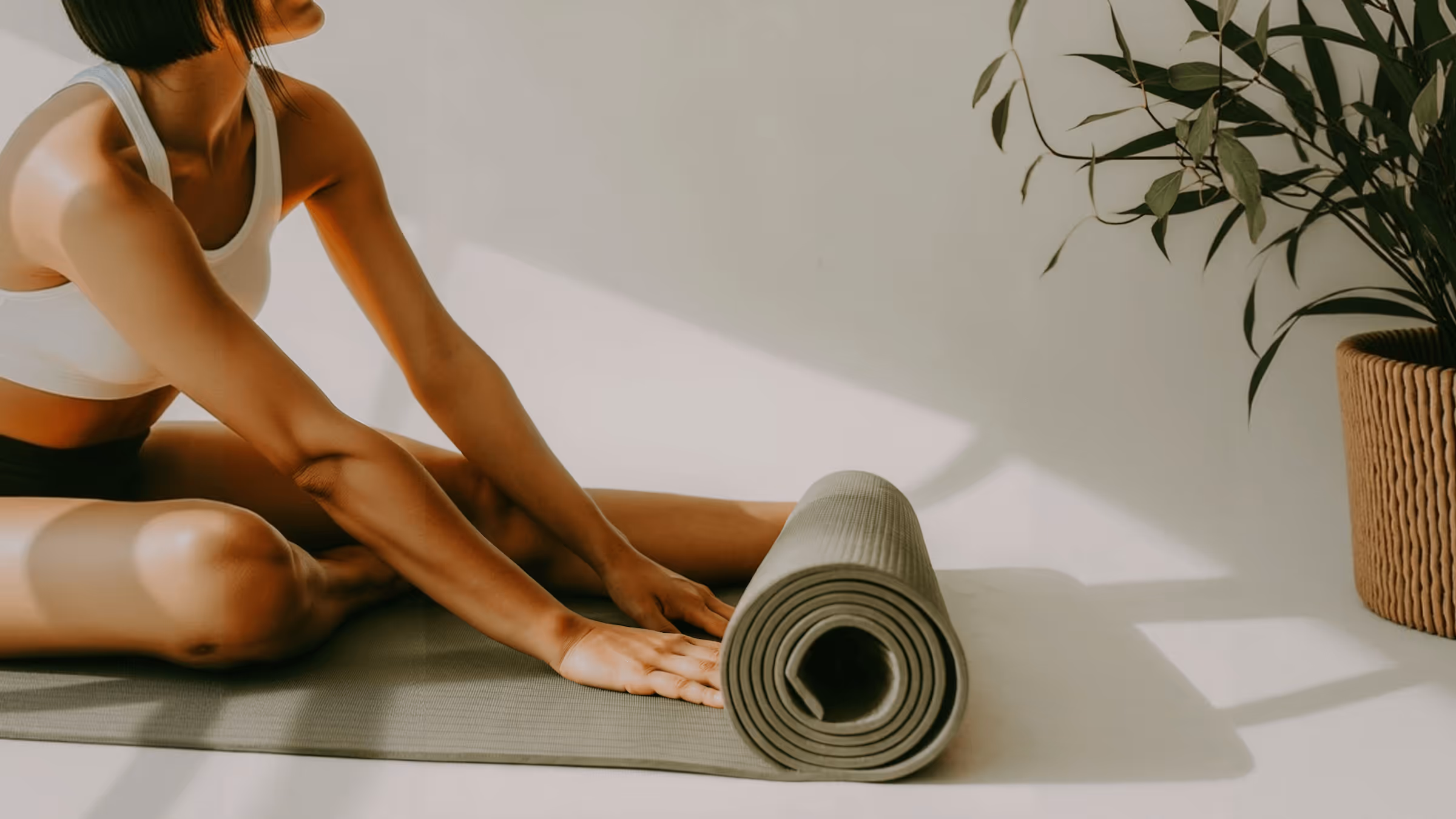 Woman in white sports bra sitting on a yoga mat, stretching forward with hands near a rolled-up mat beside her.